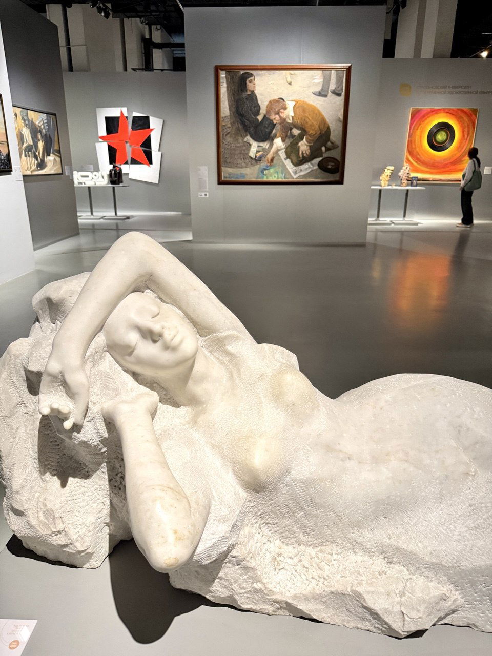 Белая мармуровая или гипсовая скульптура лежащей женщины на первом плане; на заднем плане живопись и небольшие скульптуры в зале.