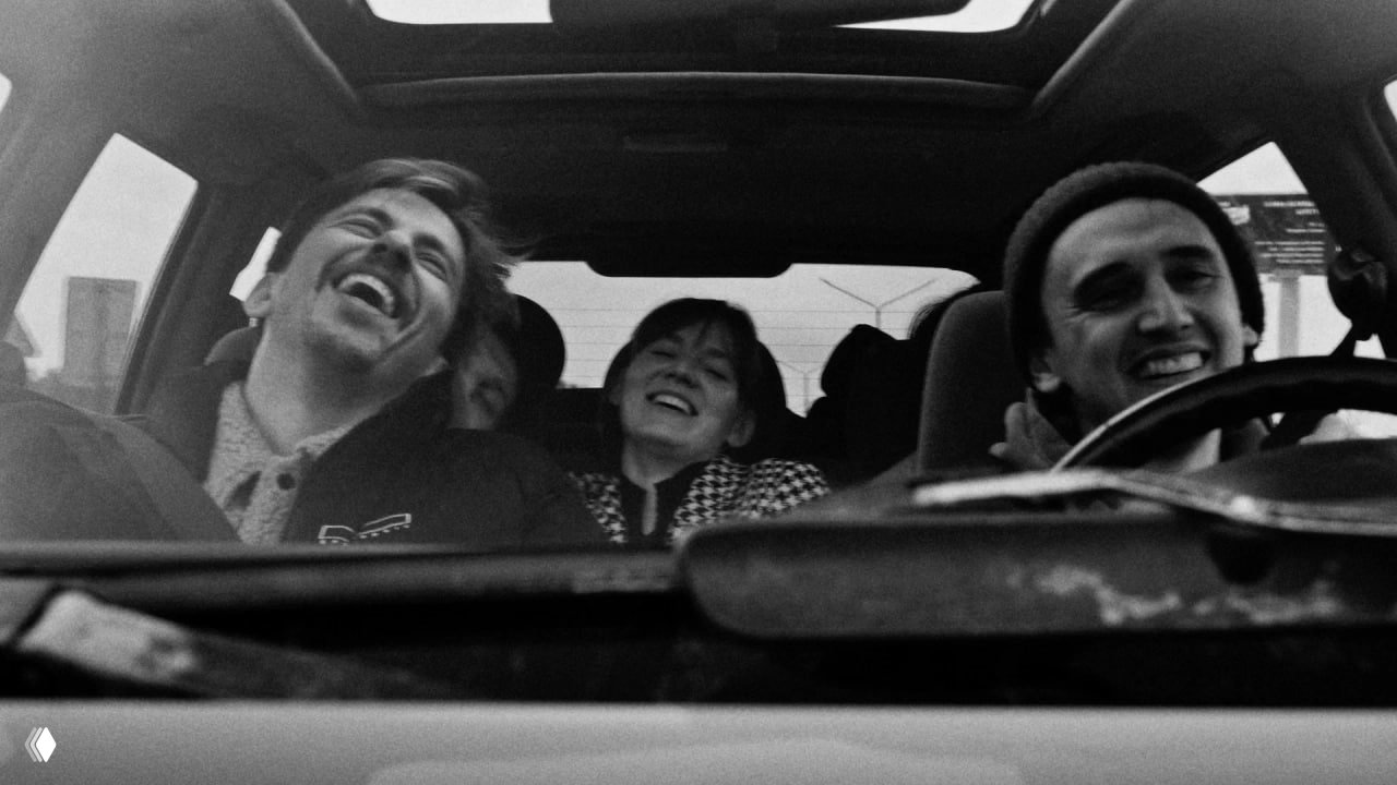 Кадр из фильма: чёрно‑белая сцена в салоне автомобиля, где трое друзей смеются и ведут непринуждённый разговор во время поездки.