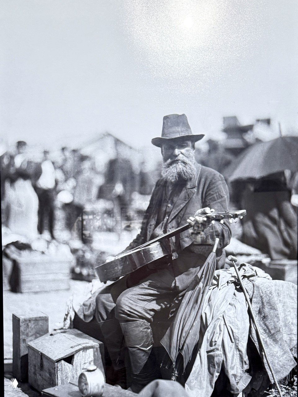 Портрет уличного музыканта на рыночной площади: бородатый мужчина с гитарой среди торговли и прохожих, типичная сцена городской хроники того времени.