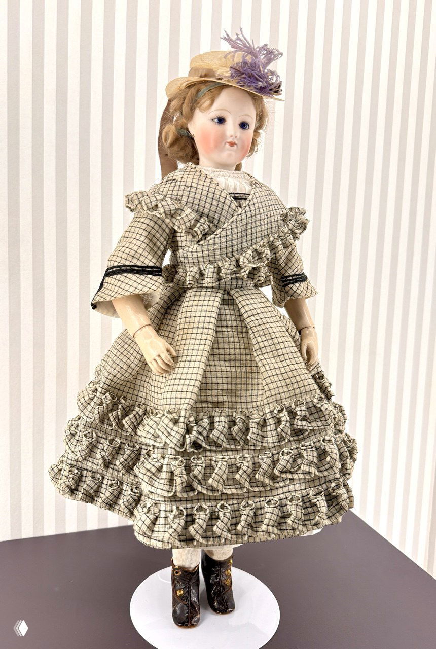 Кукла в клетчатом платье в полный рост на подставке — характерная мода конца XIX века, виден интерьер витрины с полосатым фоном.