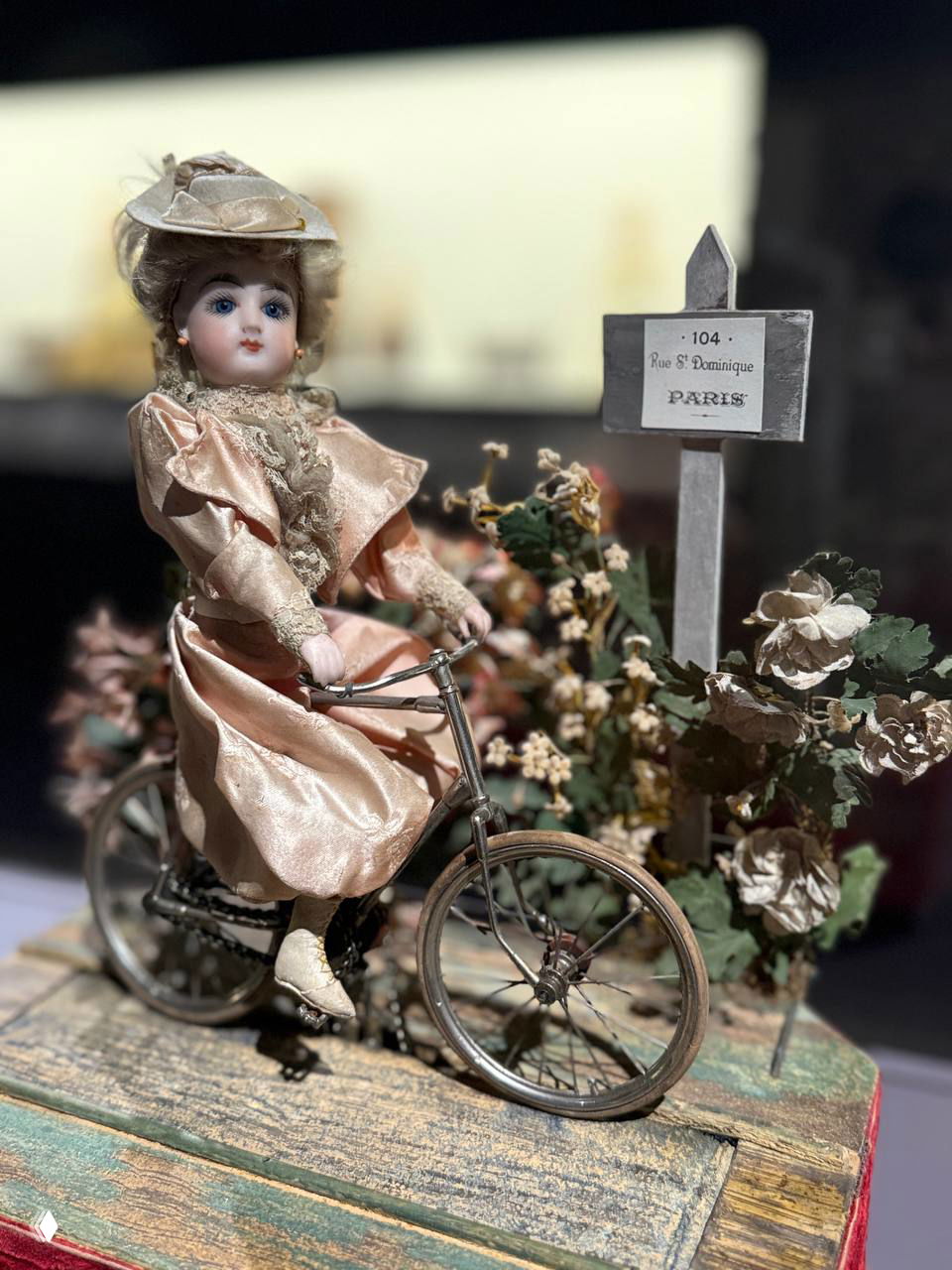 Миниатюрная кукла на велосипеде в декоративном антураже, текстуры ткани и мелкие детали реквизита хорошо видны на снимке экспозиции.