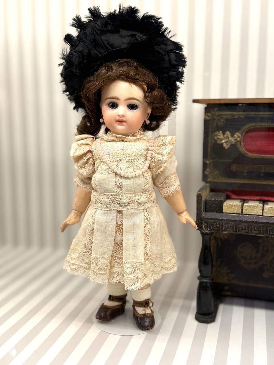 Фарфоровая кукла в светлом платье и большой чёрной шляпе рядом с антикварным комодом — акцент на аксессуарах и деталях костюма.