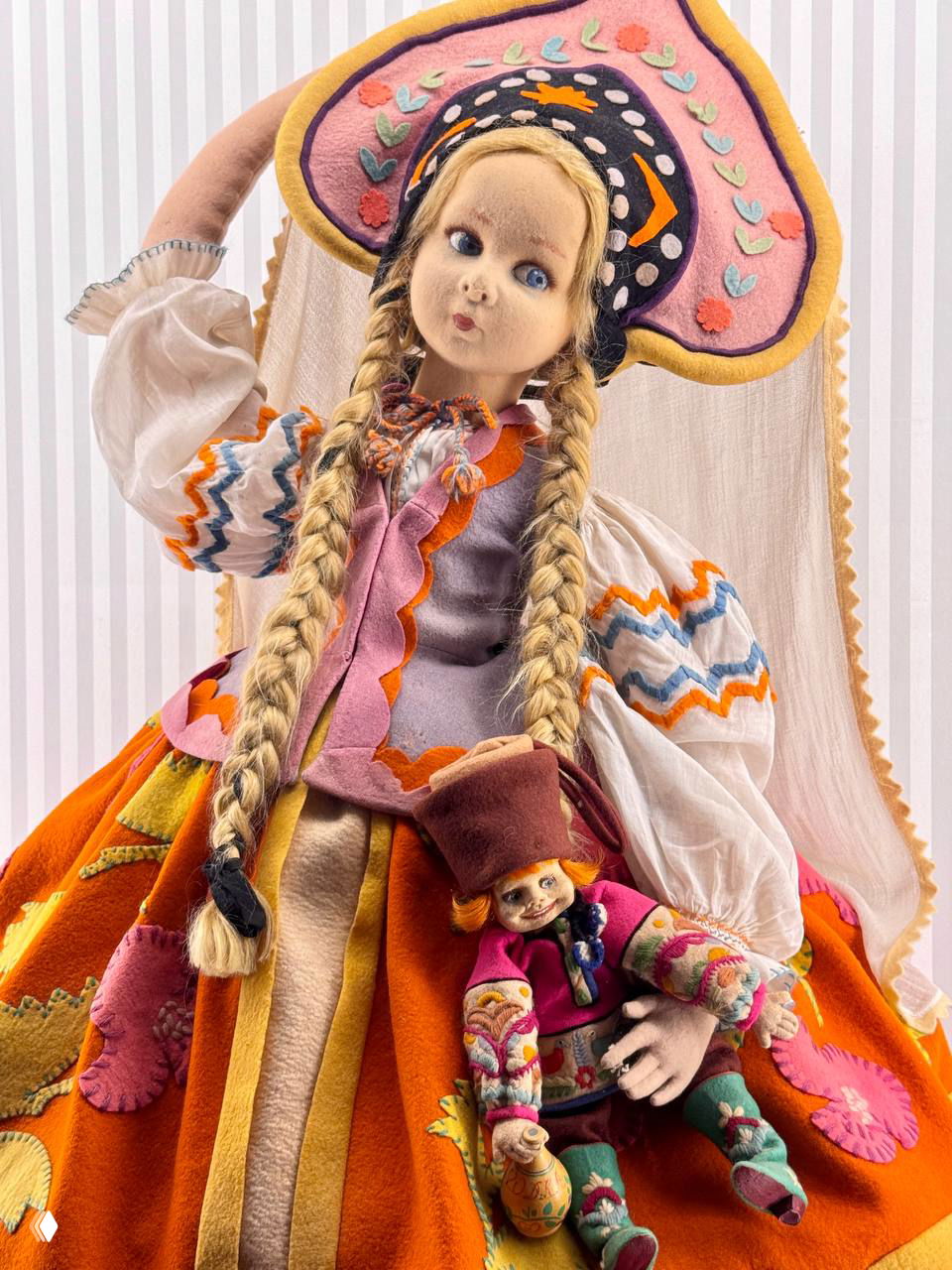 Яркая текстильная кукла в русском народном костюме с длинными косами и маленькой куколкой в руках — контраст материалов и цвета в экспозиции.