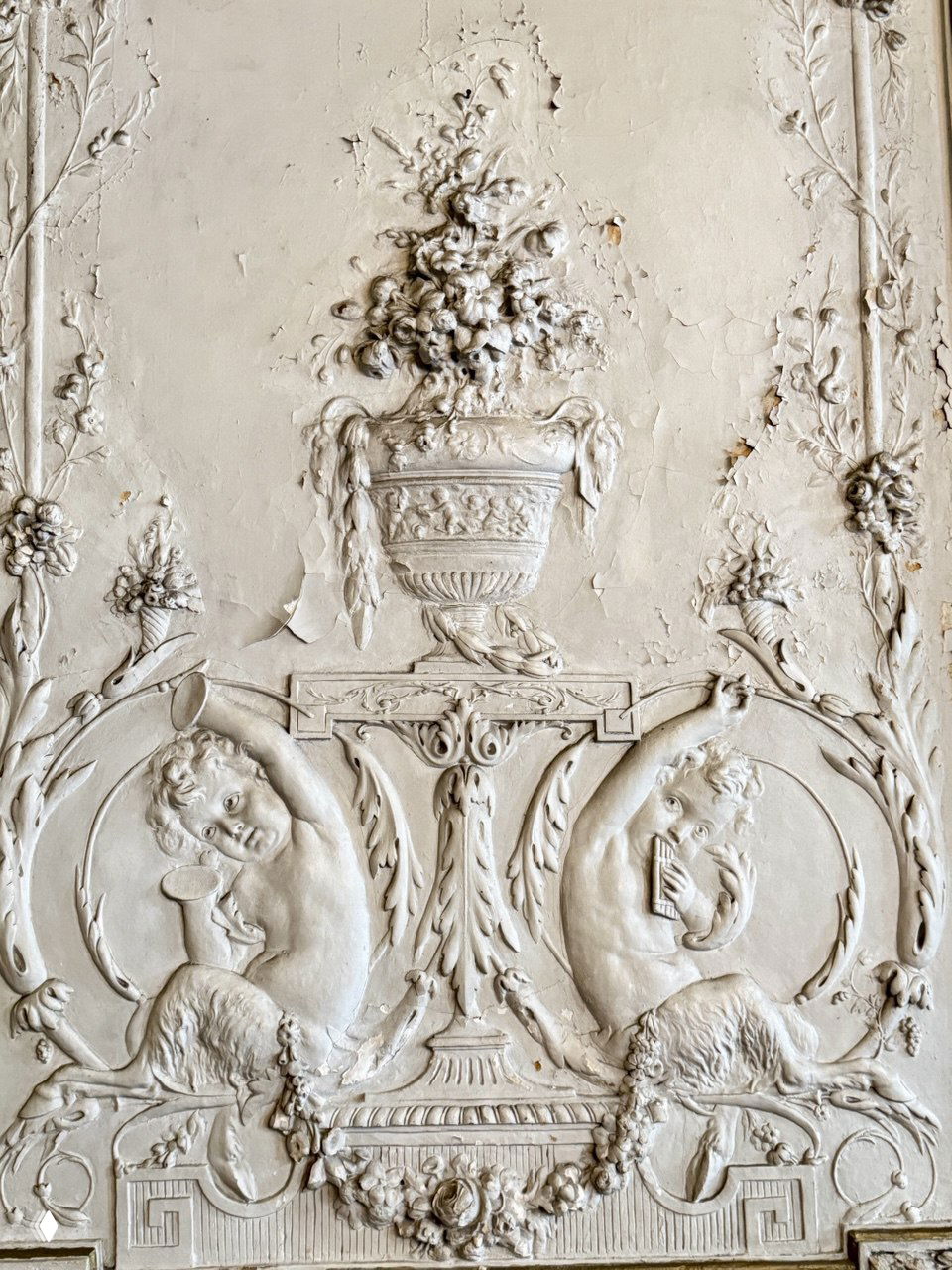Фрагмент лепнины в светлом интерьере особняка: вазон с цветами и два амура в окружении орнамента, видны трещины и следы старения.