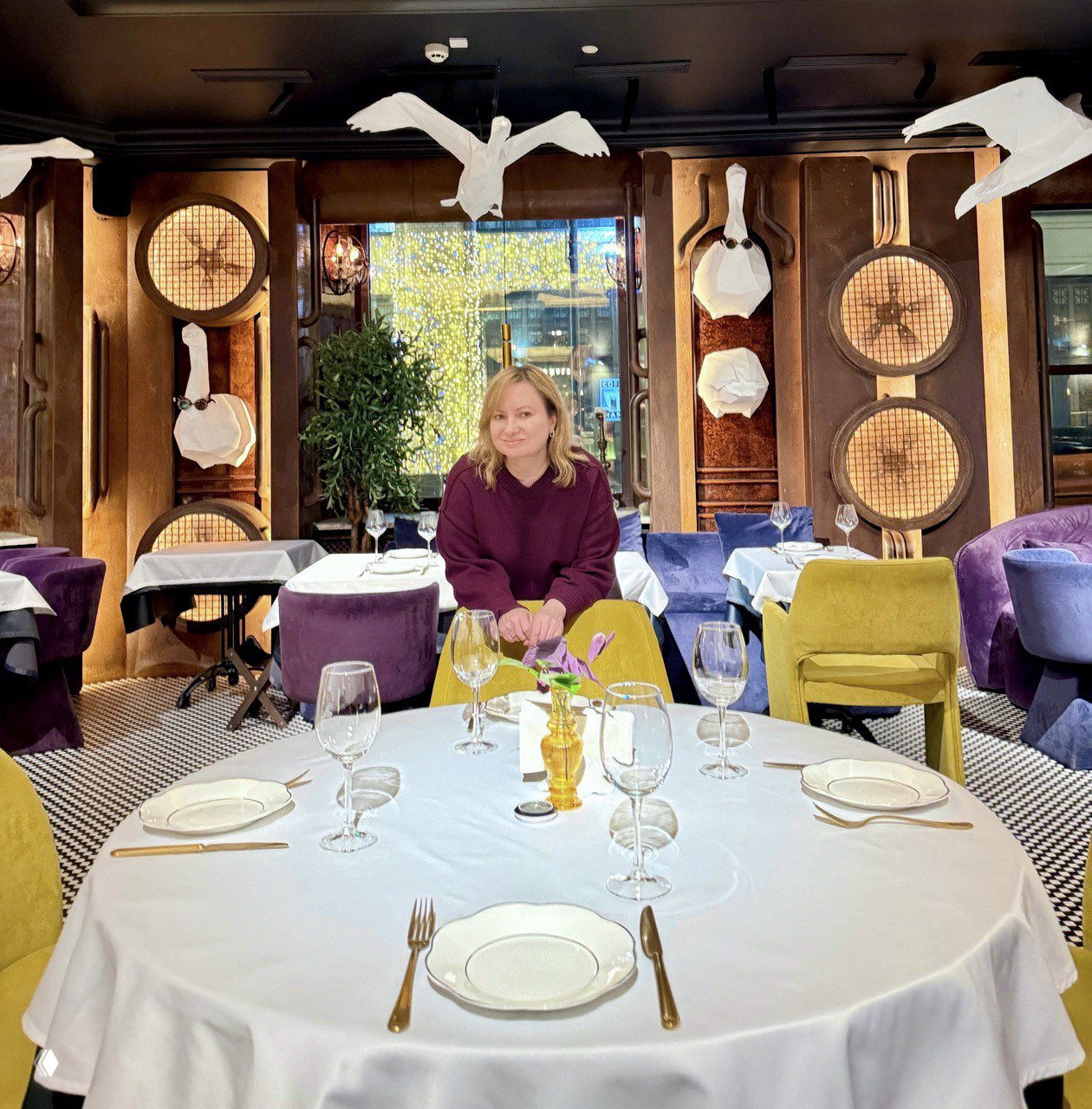 Зал ресторана Gussi с автором на переднем плане: разноцветные кресла, подвесные декоративные птицы и большие круглые элементы на стенах, придающие сценичность интерьеру.