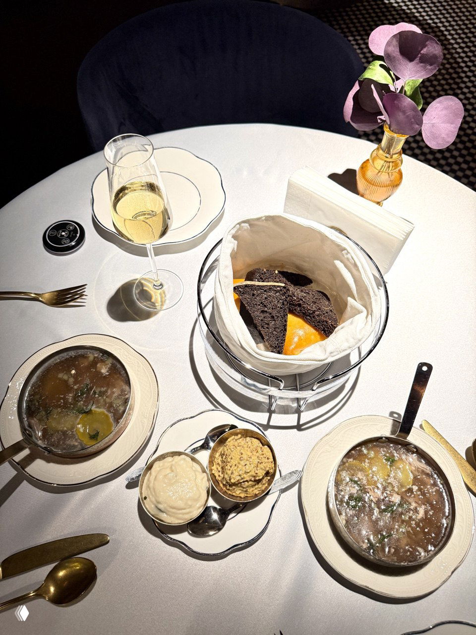 Блюда и сервировка на столе в Gussi: корзинка с хлебом, два маленьких супа, соусы и бокал шампанского на белоснежной скатерти, вид сверху на компоновку блюд.