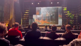 Зрители в зале смотрят показ документального фильма на большом экране; видна сцена, сидящие люди и атмосфера премьеры.