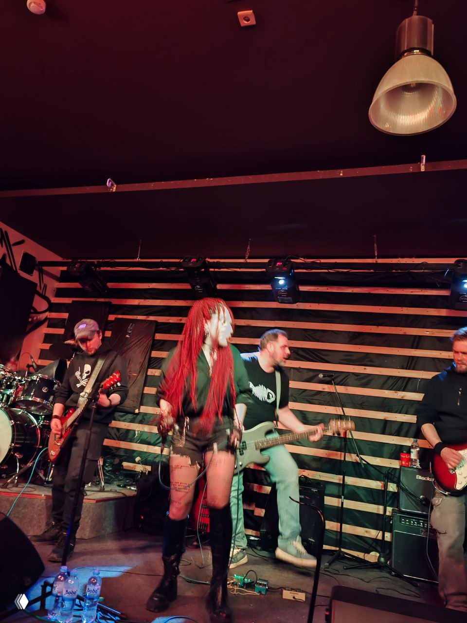 Концертный кадр: вокалистка с яркими косами и гитаристы на сцене с деревянным фоном и подвесными светильниками, момент живого выступления.