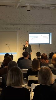 Ведущий или солист говорит с публикой, зрители сидят в рядах белых стульев, видны спины аудитории и окно с дневным светом, камерный концерт Cover Black