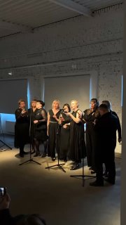 Группа певцов Cover Black под сценическим светом, несколько солистов в центре, видно белую стену и экран на заднем плане, вечернее выступление