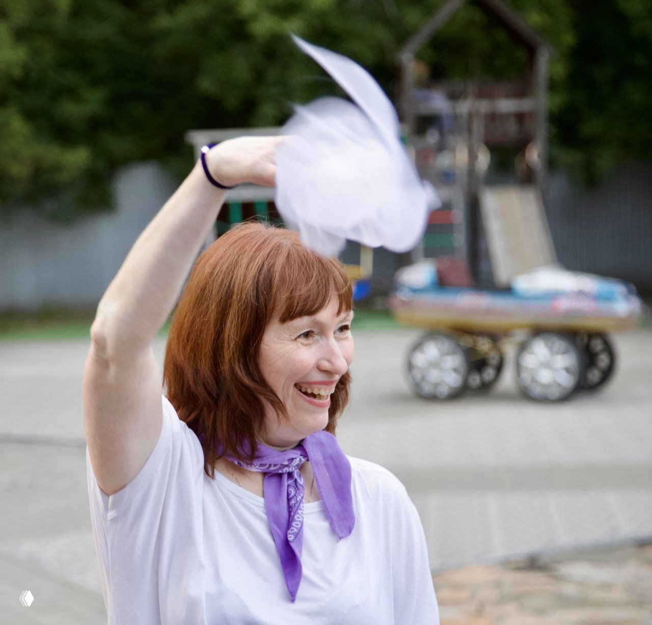 Женщина в белой футболке и сиреневом платке машет белым платком, улыбаясь на открытом пространстве, кадр передает лёгкость и радость.