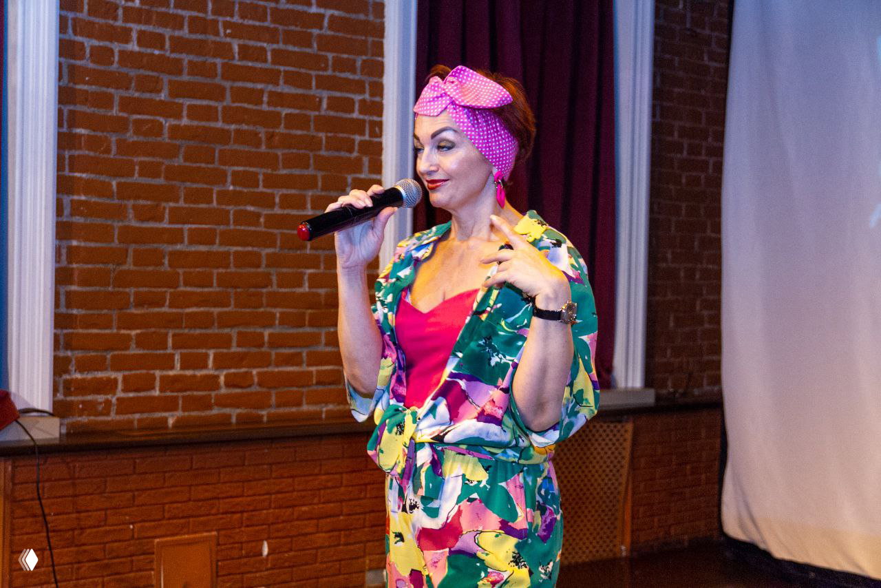 Певица в ярком образе с микрофоном на сцене — яркое цветное платье и платок на голове, сценическое выступление в интерьере клуба.