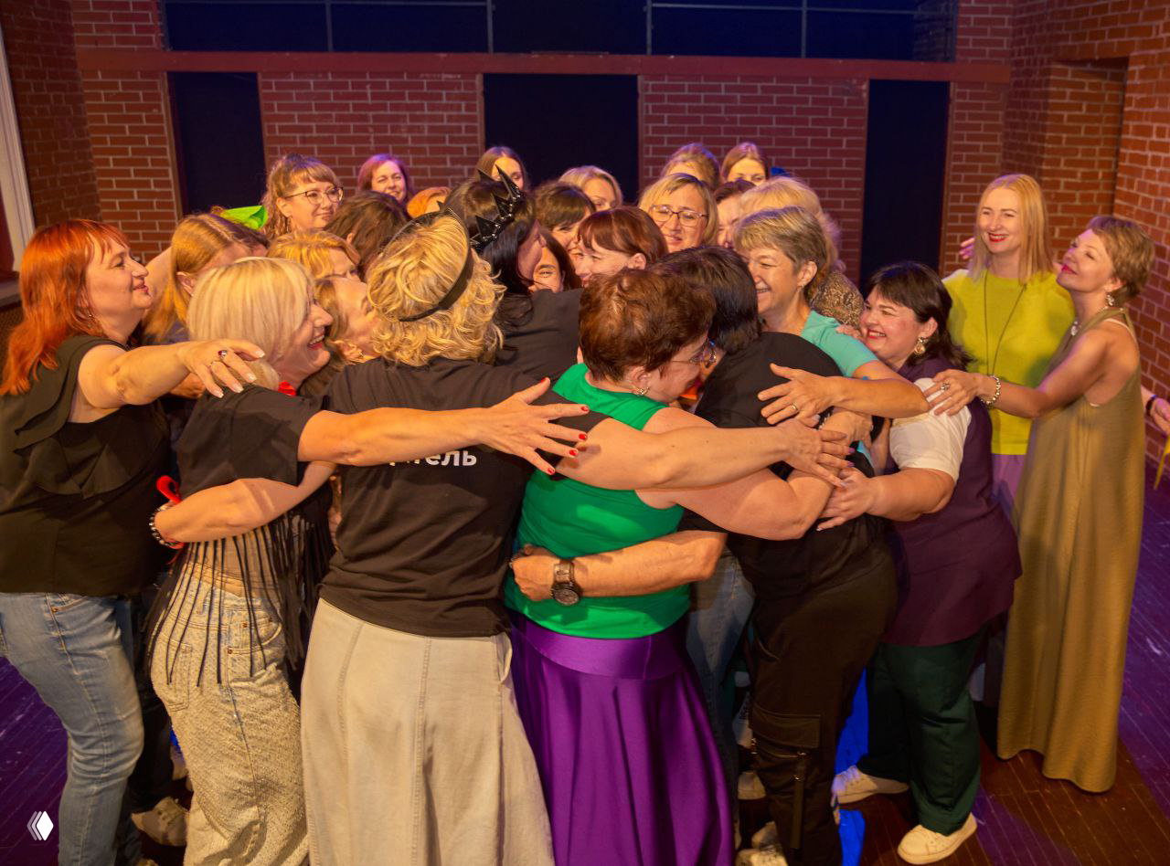 Большая группа женщин обнимается в дружеском кругу на сцене; видно много эмоций, праздник и коллективное единение.