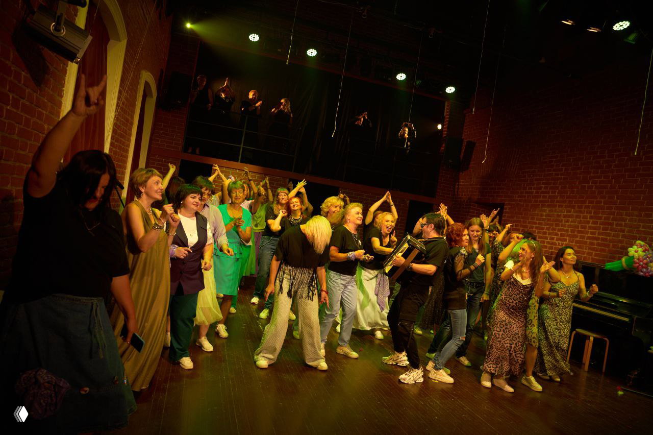 Широкий план сцены с группой женщин, аплодисментами и танцем: праздник, динамика и тёплое сценическое освещение.