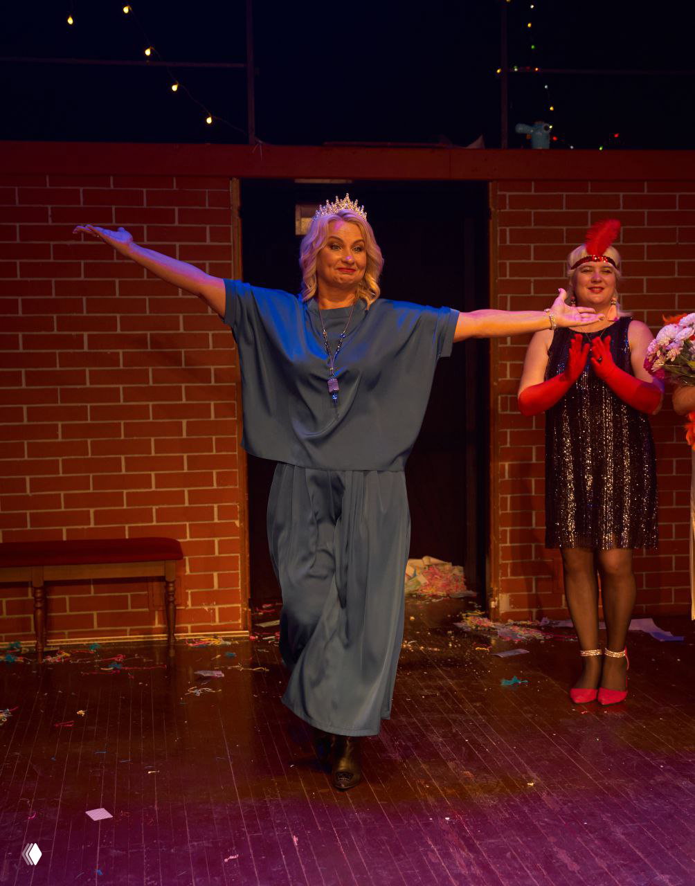 Женщина в голубом наряде идёт по сцене с разведёнными в стороны руками, вокруг — праздничный декор и аплодирующие участницы.