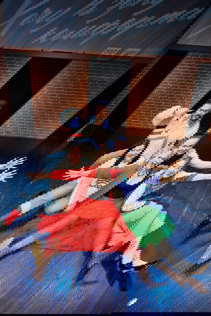 Группа артистов сидит и лежит на сцене в разноцветных костюмах, эмоциональная поза и дружеская атмосфера после номера.