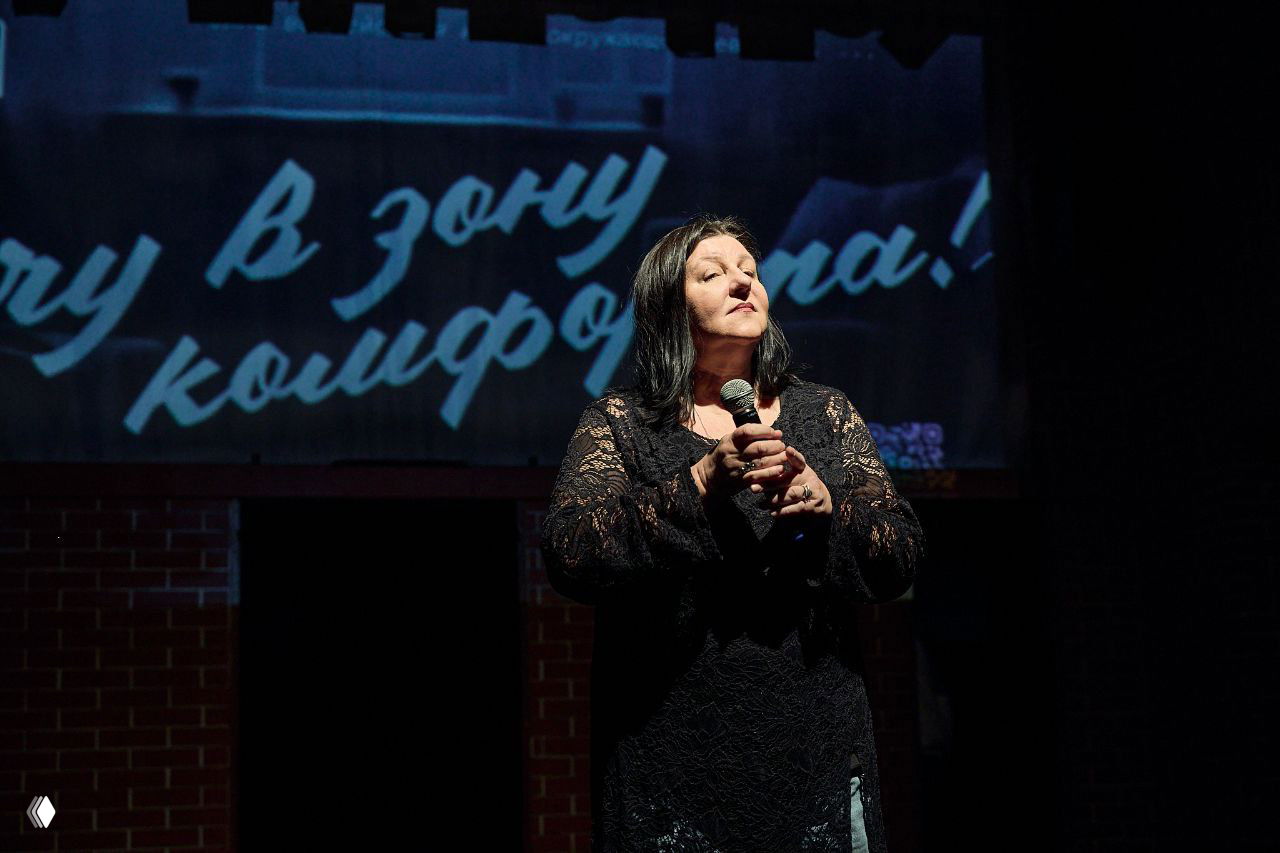 Солистка держит микрофон в руках на затемнённой сцене, крупный план с проекцией текста на заднике и театральной атмосферой.