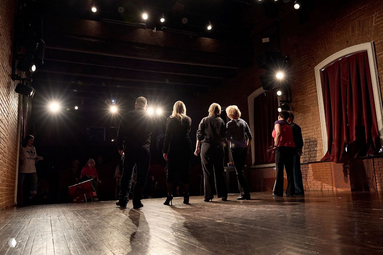 Артисты на сцене в позе друг за другом, в центре — женщина в светлой толстовке Adidas, сценическое освещение и деревянный пол.