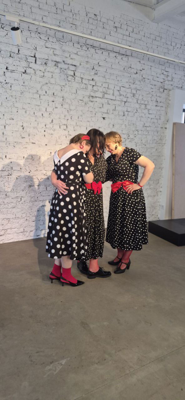 Три женщины в чёрных платьях в горошек обнимаются на сцене перед выступлением — момент репетиции у белой кирпичной стены с минималистичной сценографией.