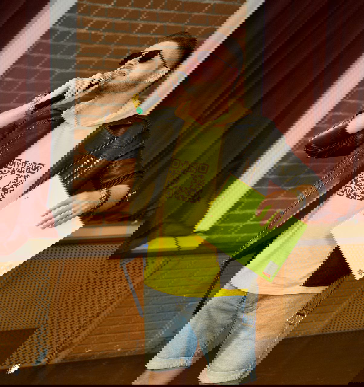 Певец в жёлтой толстовке и кожаной куртке с солнцезащитными очками держит микрофон и папку, сценический кадр в зале