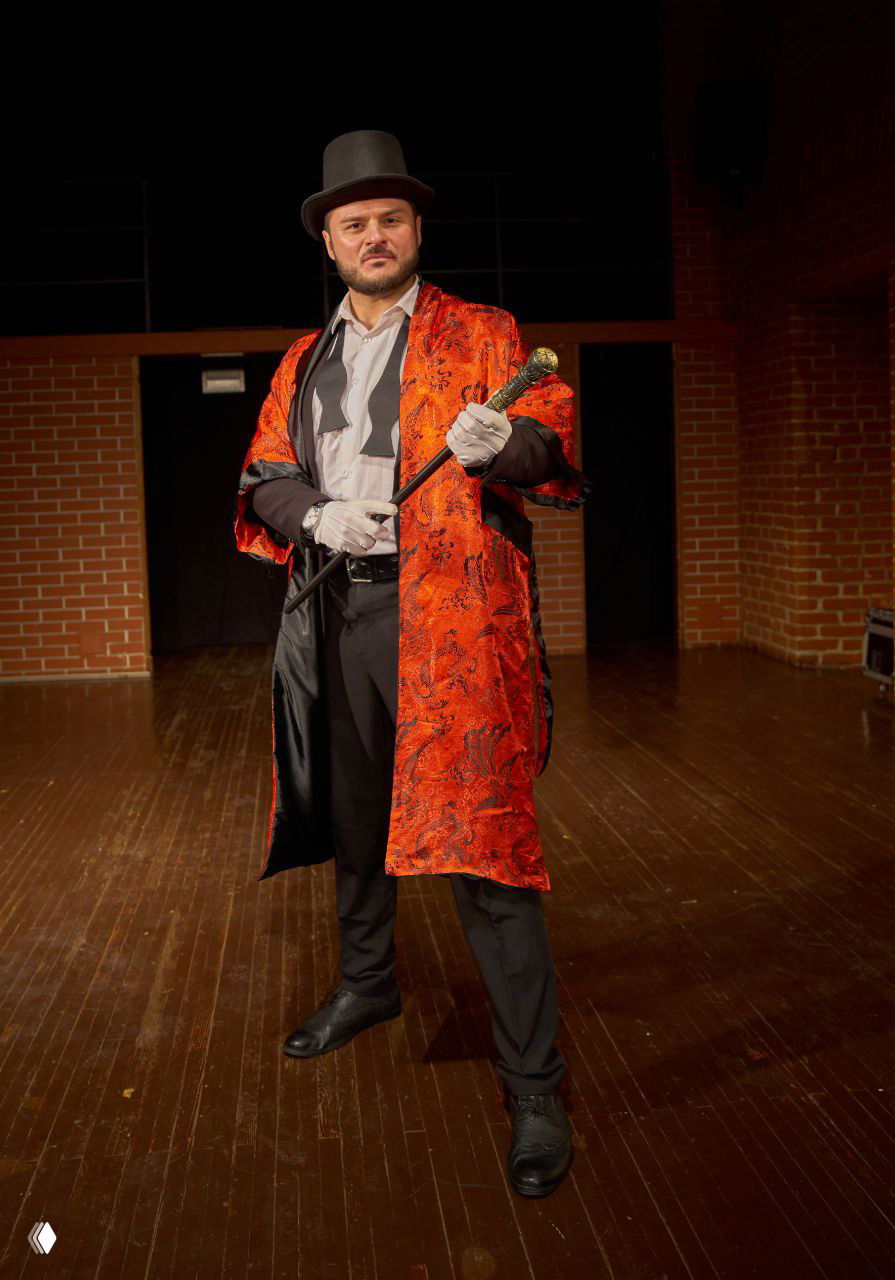 Александр Дьяконов в красном театральном плаще и цилиндре держит трость, костюмный образ на деревянной сцене с кирпичной стеной