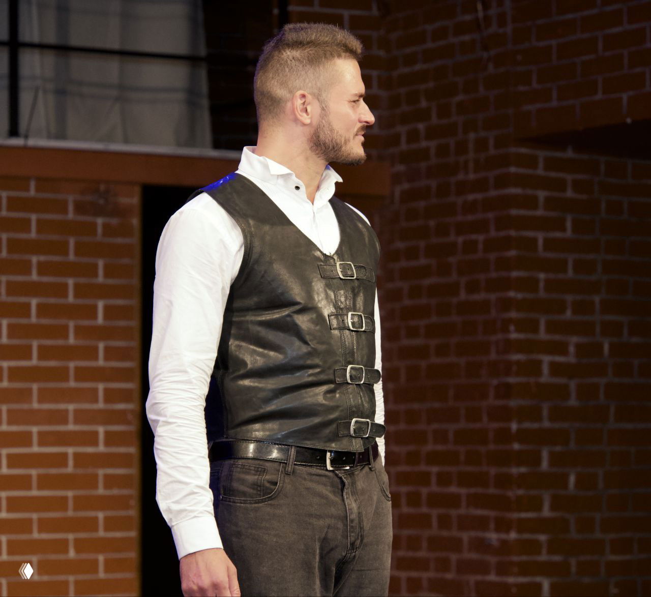 Певец в белой рубашке и чёрном кожаном жилете в профиль на сцене, спокойный драматичный портретный кадр с кирпичной стеной