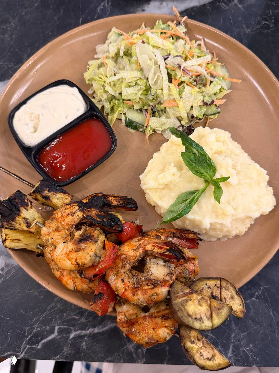 Тарелка с гриль-креветками и кусочками ананаса, баклажаном, порцией пюре и салатом; видны две ёмкости с соусами.