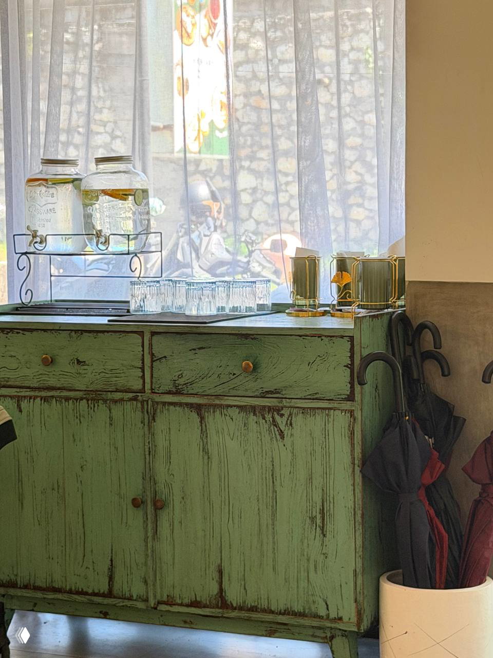 Интерьер кафе: салфетки и стеклянные емкости с водой на зелёном старинном комоде и зонты в углу, уютная деталь оформления.
