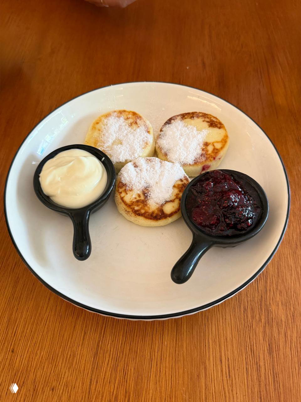 Три небольших сырника, посыпанные сахарной пудрой, с порциями сметаны и ягодного джема в маленьких соусниках на белой тарелке.