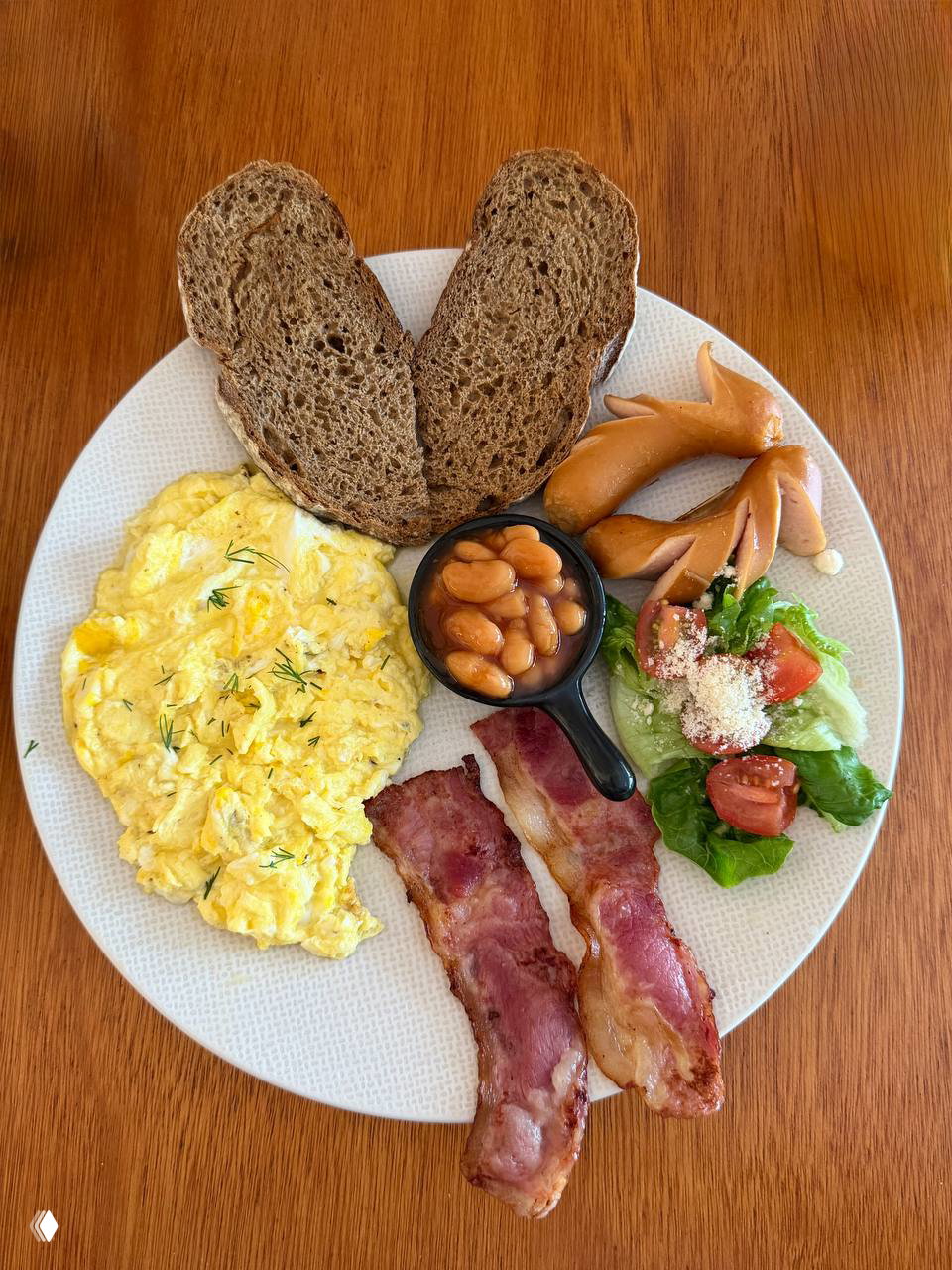 Большая тарелка завтрака: омлет, два ломтика ржаного хлеба, бекон, сосиски, фасоль в соусе и небольшой салат на белой тарелке.