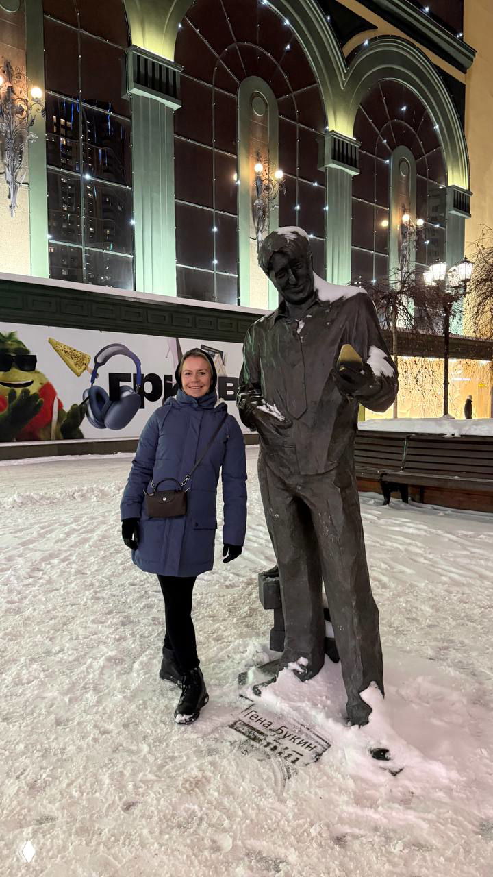 Автор поста фотографируется рядом со скульптурой Геннадия Букина вечером на заснеженной площади: памятник и подсветка торгового центра.