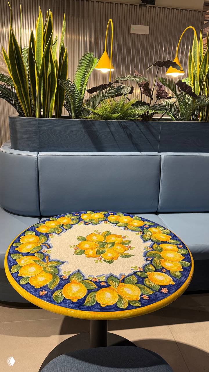 Круглый стол с лимонным орнаментом на переднем плане, диван, растения и лампы — лаунж-коворкинг CX Napoli Centrale вблизи вокзала.