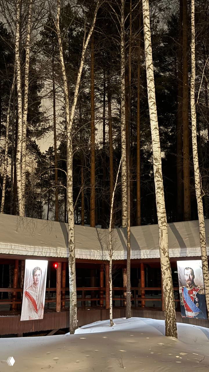 Ночная фотка Ганиной Ямы: деревянная галерея среди берёз и плакаты с портретами царской семьи в окружении снега и сосен.