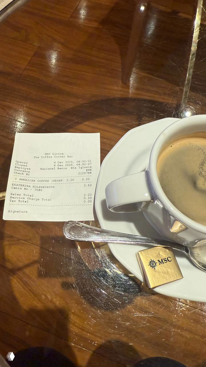 Чашка кофе на блюдце и чек на столе кафе лайнера, рядом маленький квадратик шоколада с логотипом MSC — повседневный момент на круизе.