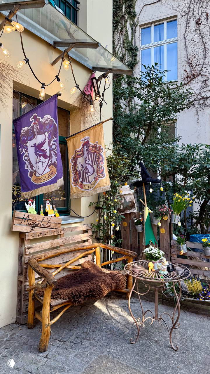 Уютный внутренний дворик рядом с витриной: флаги факультетов Хогвартса, деревянная лавочка и декор перед входом в магазин.