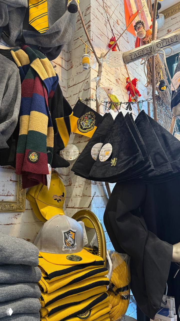Полки с тематическими шарфами, колпаками и атрибутикой факультетов: вешалки с тёмными накидками и жёлтыми акцентами товаров.