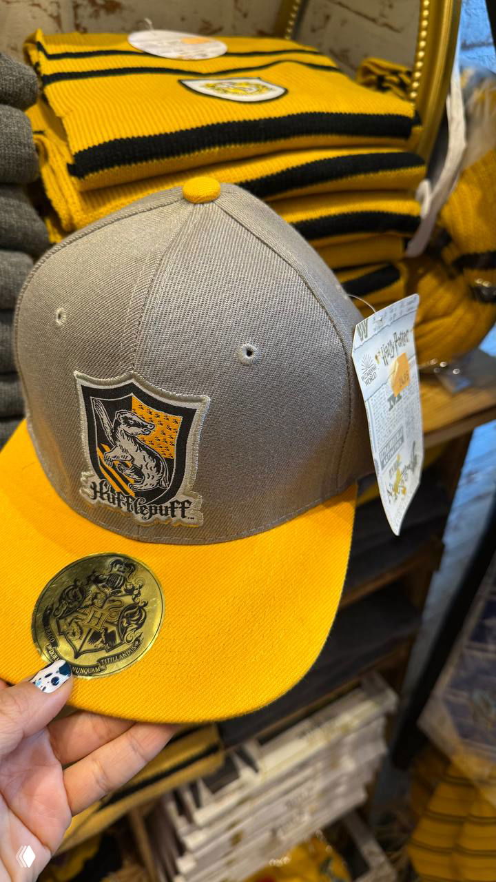 Крупный план кепки Hufflepuff жёлто‑серого цвета на фоне стопки свитеров и брендированных товаров внутри магазина.