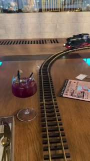 Крупный план рельсов на столе внутри ресторана и модель‑поезд в движении, рядом бокал с напитком — способ подачи заказов по столам.