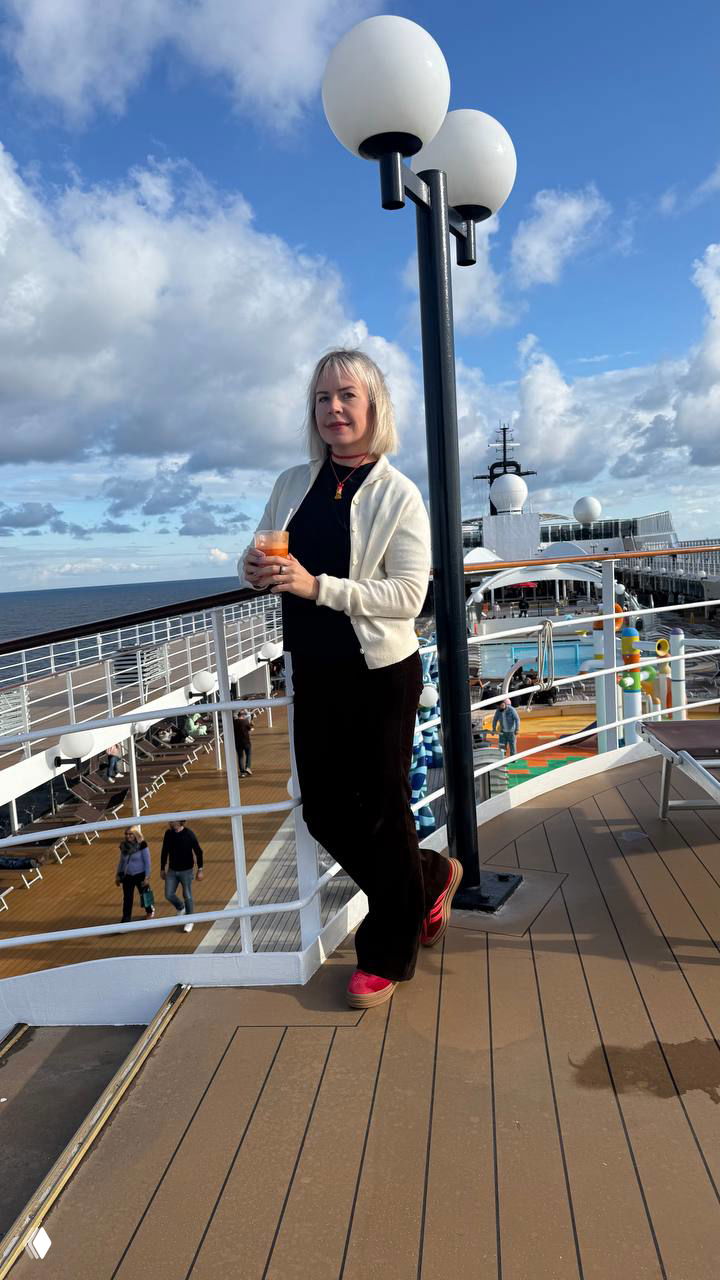 Автор стоит на верхней палубе лайнера с напитком в руках, открытый вид на палубу и море, ясное небо и прогулочная зона на заднем плане.