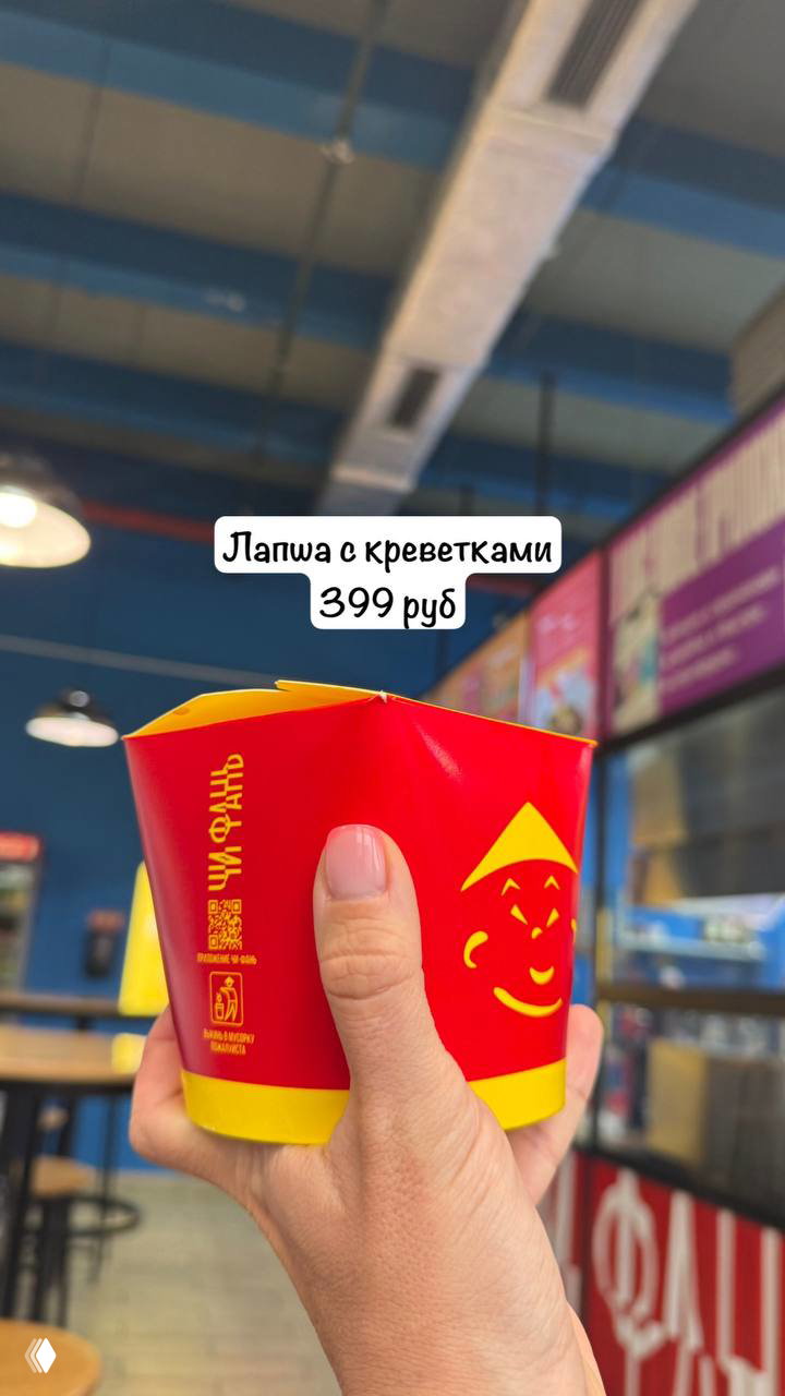 Рука держит упаковку с китайской лапшой вок, на фоне интерьер фудкорта; этикетка показывает цену 399 руб для лапши с креветками.