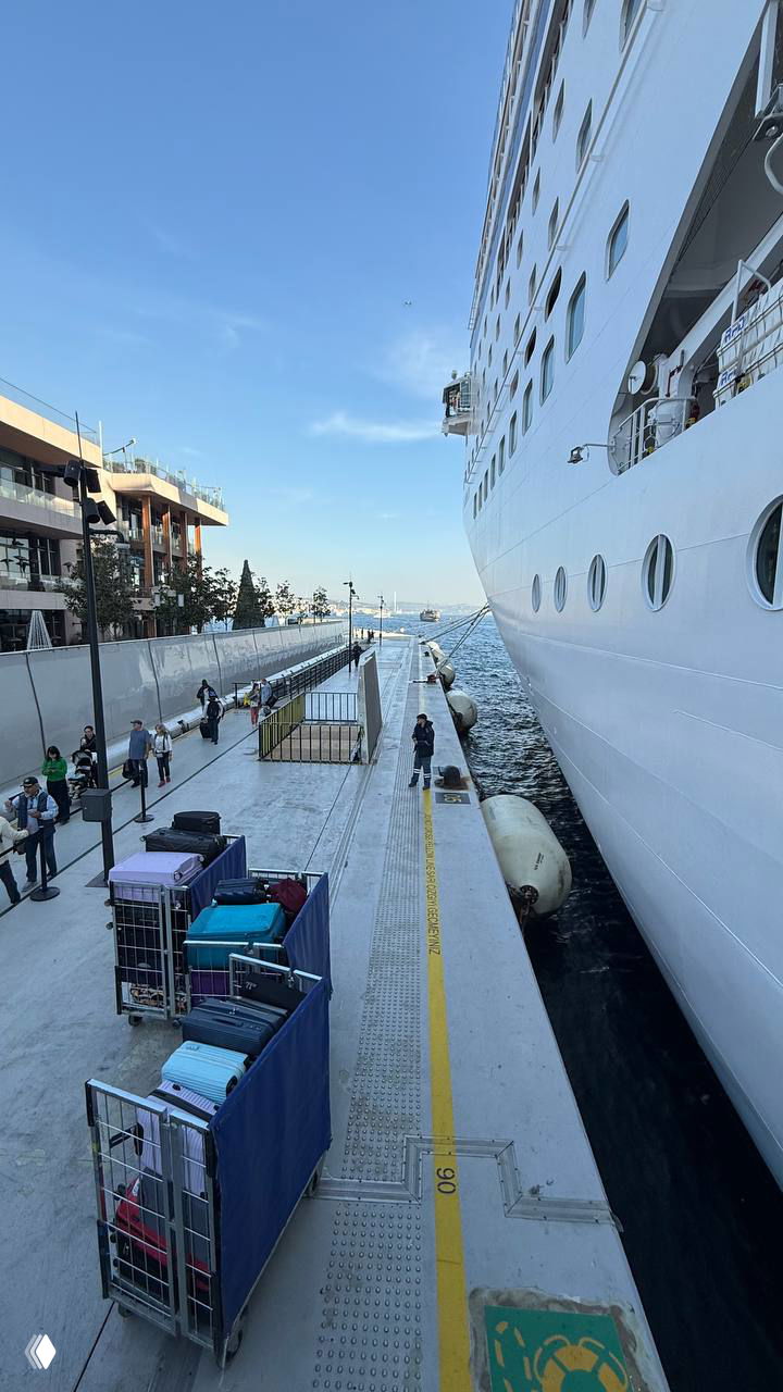 Пирс в порту рядом с огромным круизным лайнером: тележки с багажом на причале и сотрудники порта — сцена высадки/погрузки.