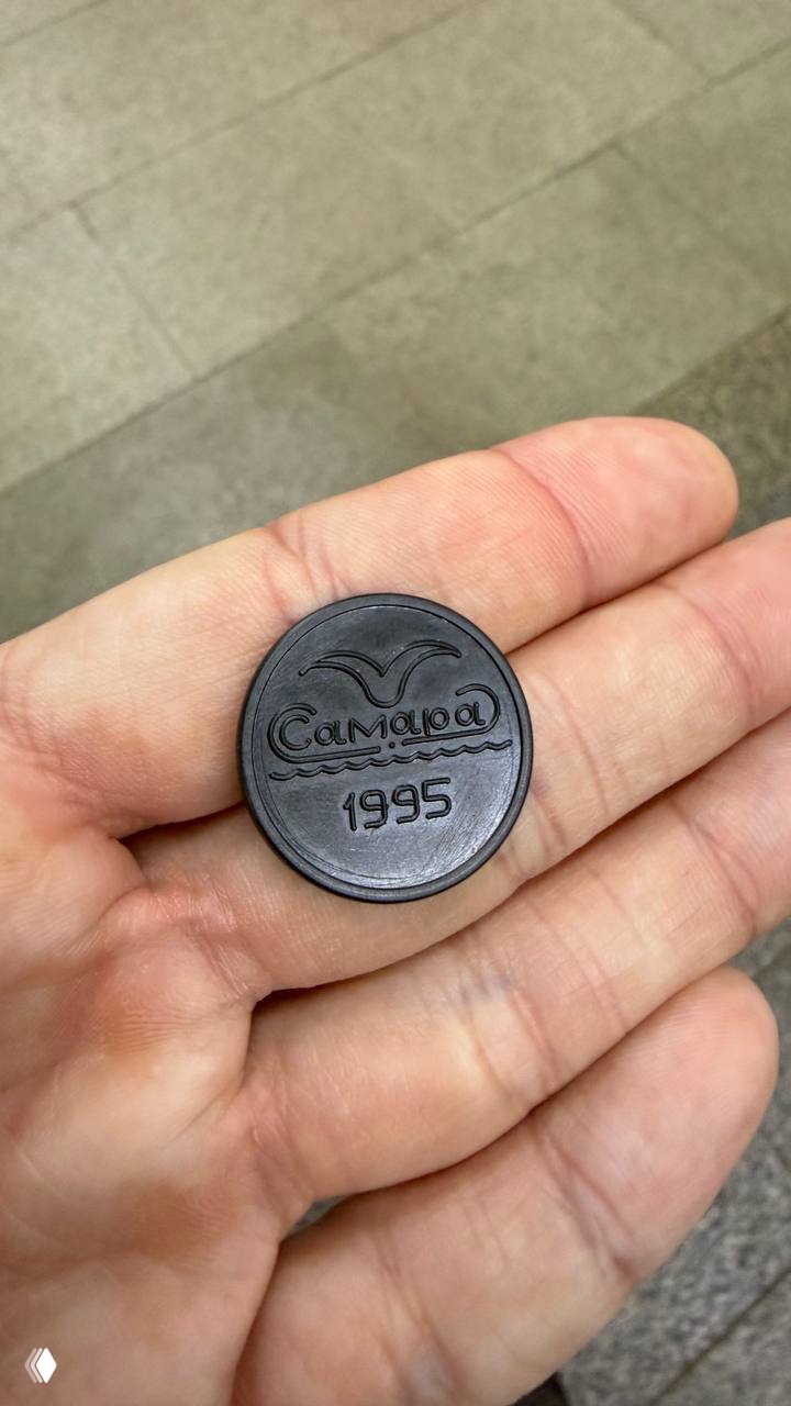 Рука держит чёрный пластиковый жетон самарского метро с надписью «Самара 1995» на фоне пола станции — крупный план