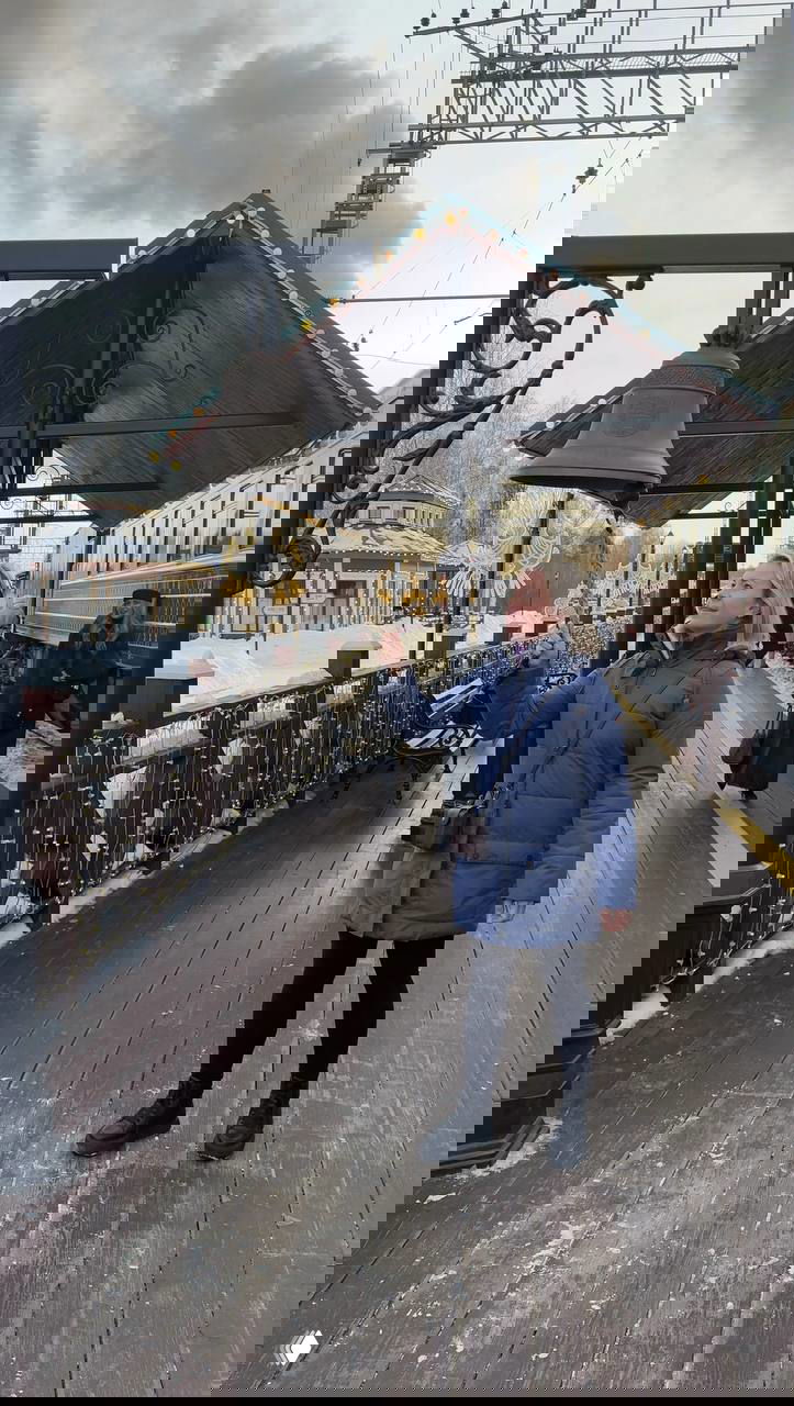 Женщина в тёплой куртке звонит в старинный колокол на деревянной платформе ретро-станции — рядом прицеплен паровоз и видна железнодорожная линия.