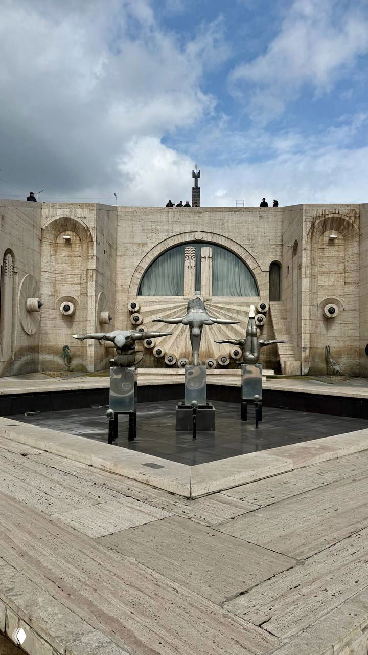 Фонтан и архитектурный фрагмент Каскада в Ереване: каменные ступени, скульптуры и прохожие на вершине на фоне неба.