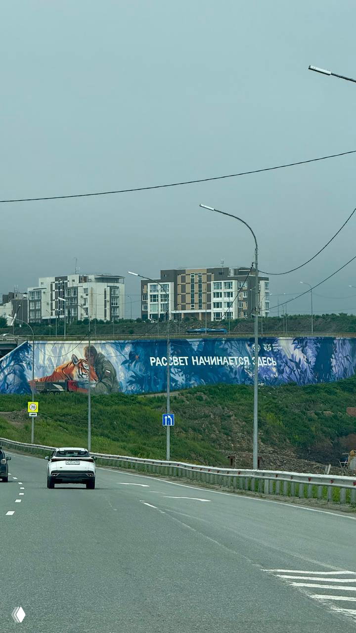Дорога к городу с панорамой жилых домов на холме и большим муралом с надписью «РАССВЕТ НАЧИНАЕТСЯ ЗДЕСЬ», машины на асфальте впереди.