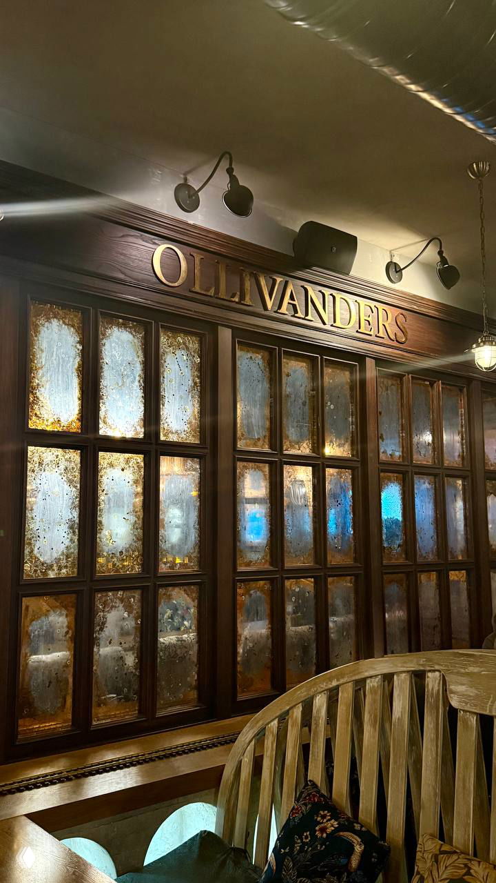 Деталь интерьера с надписью OLLIVANDERS над тёмной деревянной витриной — отсылка к лавке волшебных палочек, состаренное стекло и резное дерево.