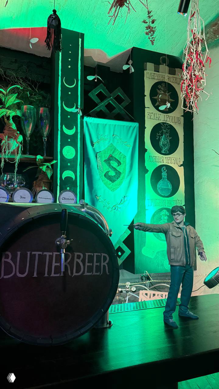 Сцена с бочкой «butterbeer», сценическими декорациями и фигурками — место для мероприятий и фотозон внутри бара-музея, подсвеченное зелёными тонами.