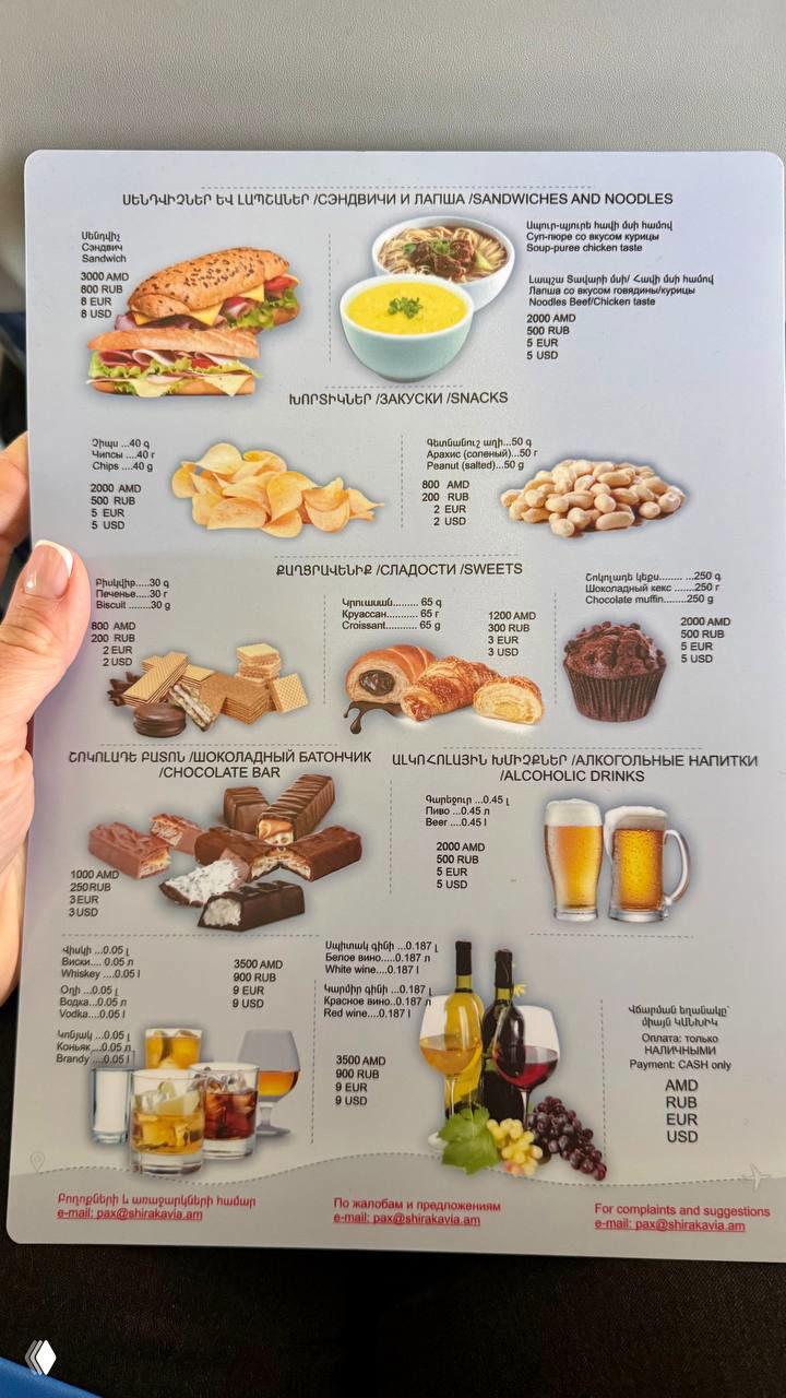 Лист меню авиакомпании с разделом «закуски» и ценами в AMD/RUB/USD — фотография в руке пассажира, видно ассортимент снеков и сладостей.