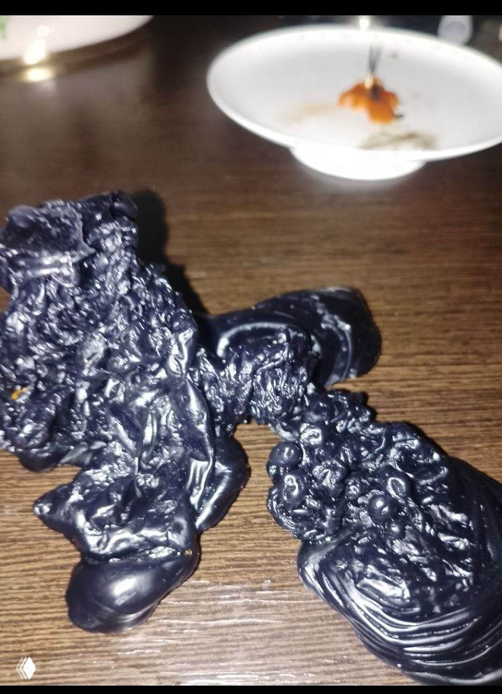 Чёрные куски застывшего воска на деревянной поверхности, вдалеке чашка с остатками воска; композиция показывает результат отливки и её объём.