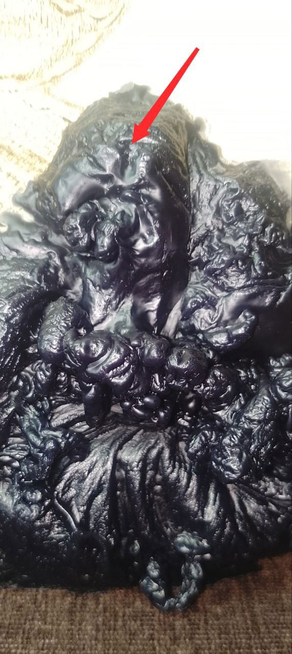 Отливка чёрного воска на светлом фоне; видна фигура с лицевой стороной, на фото отмечено красной стрелкой место, где проглядывают черты лица.